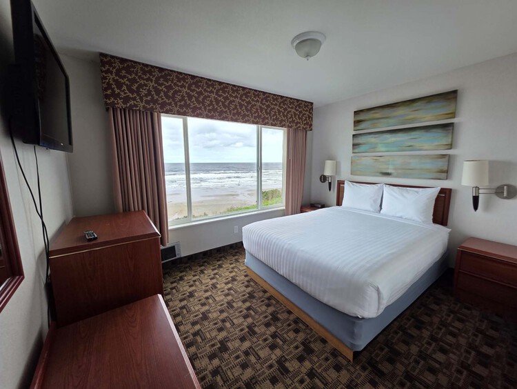 A guest room with a queen bed, a TV, and a large window with a view of the beach